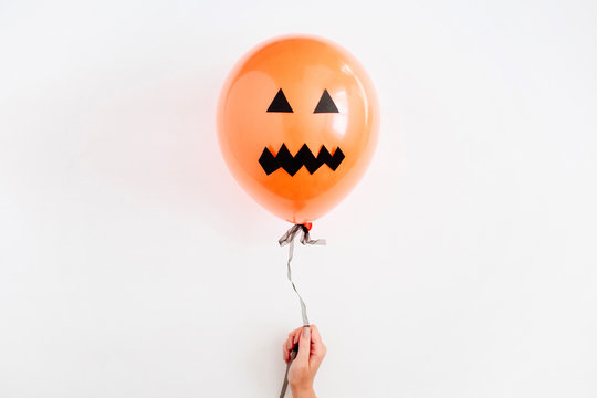Halloween minimal concept. One orange balloon with scary face in girl's hand on white background. Flat lay, top view.
