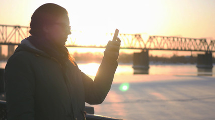 Fototapeta premium Young attractive girl is walking along the waterfront on the background of a railway bridge and a beautiful sunset, holding a phone in hands, writes a message.