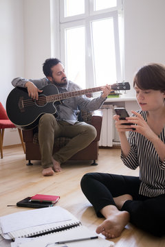 A young man is rehearsing on a bass guitar while the girlfriend is checking her smart phone