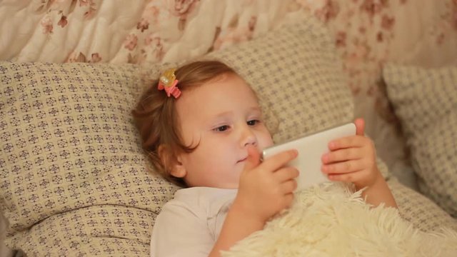 Child Looks cartoons and plays downloaded application on a smart phone close-up. A girl lies in bed and looks at the white phone screen.