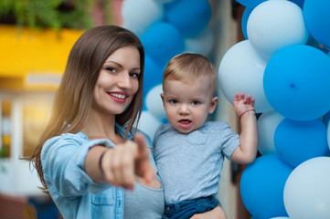 Portrait of young beautiful mother with her year-old baby boy. Woman point the finger in front of her on the camera on balloons background
