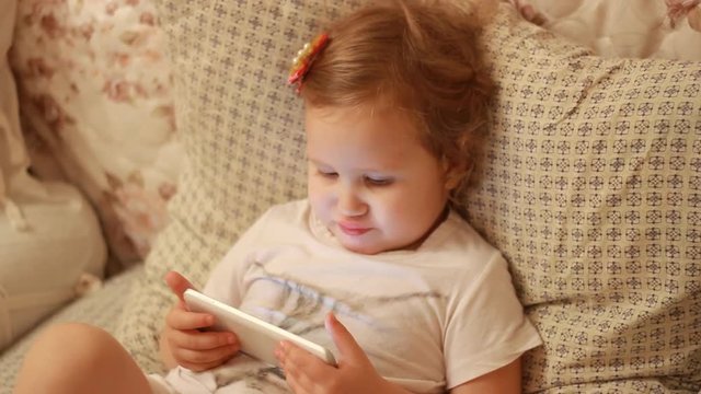 Child Looks cartoons and plays downloaded application on a smart phone close-up. A girl lies in bed and looks at the phone screen.