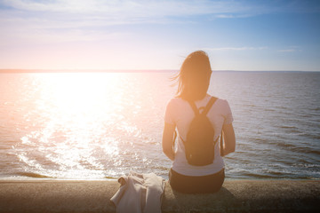 Girl sits on the stone embankment of coast sea and looks at the sun reflects on the surface of the water. In the backlight sunbeam light. Concept theme: travel, meditation, contemplation, relax, quiet