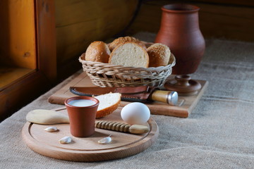 A cup of milk  and a wicker basket with bread. A clay cup with milk and a wicker basket with bread are on the table near the window in the village house.