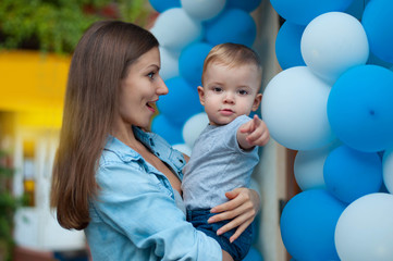 young beautiful mother in shock looks at her little boy who points a finger in front of him on the camera on balloons background
