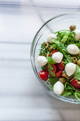 Fresh salad with tomatoes, green olives and mozzarella in glass bowl on marble table. Selective focus