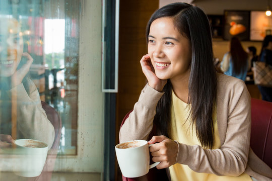 Asian woman sitting next to glass window inside coffee shop with cup of hot coffee in left hand