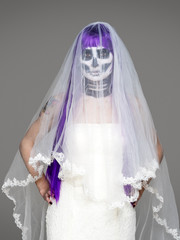 Portrait of woman looks at the camera with terrifying halloween skeleton makeup and purple wig bridal veil, wedding dress over gray background. Black wedding