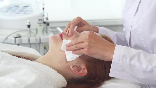 Female cosmetologist wiping her client's face with napkins. Beautician preparing pretty caucasian girl for procedures. Attractive young woman lying against background of some equipment