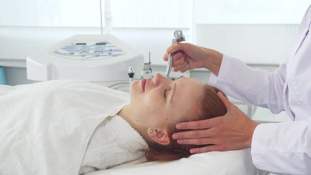 Cosmetologist using some equipment on client's face. Pretty caucasian girl lying on the couch with closed eyes. Attractive blond woman getting anti-aging procedures