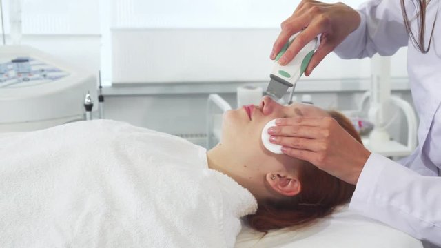 Cosmetologist in white coat using ultrasonic machine for client's face. Attractive young woman getting facial peeling. Female beautician holding handle of the peeler in one hand and cotton sponge in