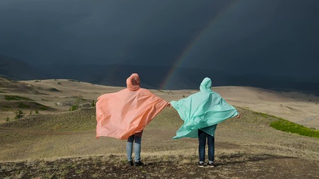 friends holding hands stand on a background of a mountain landscape with a rainbow. are dressed in raincoats. 20s 4k.