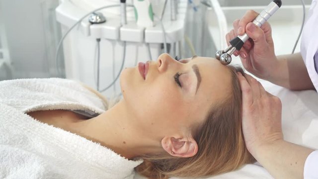 Cosmetologist applying iontophoresis on client's face. Female hands moving ball head of machine along girl's forehead. Pretty caucasian woman getting physiotherapeutic treatment for her facial skin
