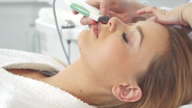 Female cosmetologist using ultrasonic scrubber on client's face. Close up of pretty caucasian woman getting deep peeling of her skin. Professional beautician moving the head of machine along girls