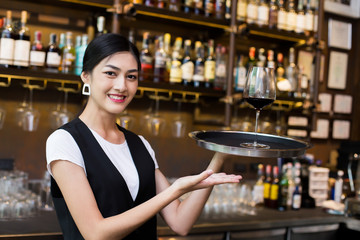 Young Asian Woman Waiter Restaurant Catering Service Happy Emotion. Woman Waiter Present Red-Wine for Customer at Bar. Woman with Wine in Bar Concept.
