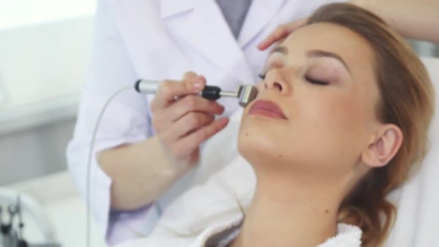 Professional beautician using iontophoresis roller for client's face. Close up of female cosmetologist moving the head of machine along woman's cheek. Pretty caucasian girl closing her eyes on beauty