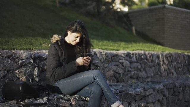 Young woman wearing jeans and a vest is sitting on a stone bench in a park, smiling and texting from her smartphone. Locked down real time establishing shot