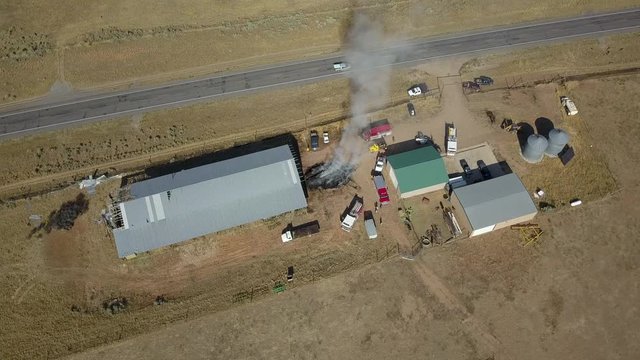 Aerial rural farm barn burning fire trucks circle. horse and cattle ranch buildings threatened by hay feed fire. Local volunteer fire fighters respond to control the blaze and minimize damage.