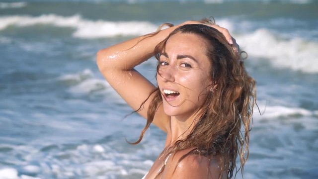 Young beautiful sexy tanned girl in bikini with curly hair sitting on the beach at sunset by the sea. She smiled enigmatically, stroked herself and. Slow motion. 120fps