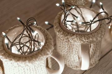 Ceramic stylish cups in sweaters and christmas retro garland on bokeh lights background. Shallow depth of field. Tinted photo.