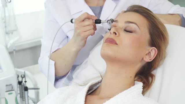 Female cosmetologist moving iontophoresis roller along client's forehead. Professional beautician performing galvanic facial treatment. Pretty blond woman getting anti-aging procedures for her skin