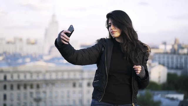 Young woman with long hair wearing black clothes is standing on a roof of a building in a city and taking a selfie. Locked down real time medium shot