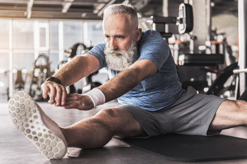 Serious old bearded man is enjoying exercises in fitness center