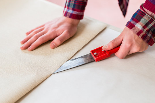 Close-up of the hands of a man cutting with a knife a new beige wallpaper sheet during home remodeling