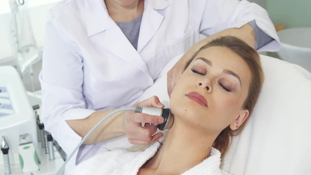 Female cosmetologist making facial massage with special equipment. Professional beautician moving the massaging kit along woman's chin and neck. Pretty caucasian girl sitting with closed eyes facing