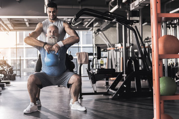 Fototapeta na wymiar Concentrated old male having workout in gym with young instructor