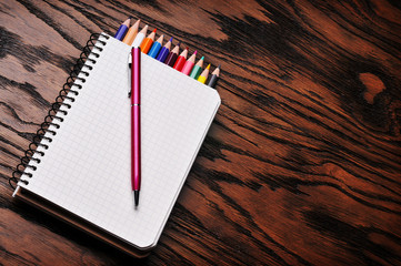 An opened notebook with pencils and a pen on the wooden background