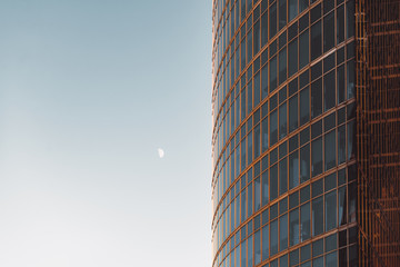 Obraz premium Side view of modern skyscraper teal and orange glassy facade with blue sky and moon on other side, offices overlooked behind windows, 
