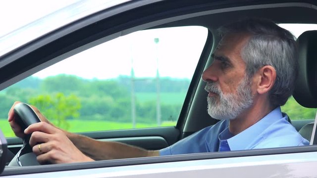 Senior man sits in the car and complains on traffic situation - closeup shot from side - countryside in the background 