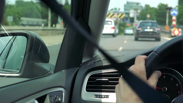 Senior man drives a car - closeup of hand on the steering wheel and speedometer - shot over seat belt - other cars on the road in the background 