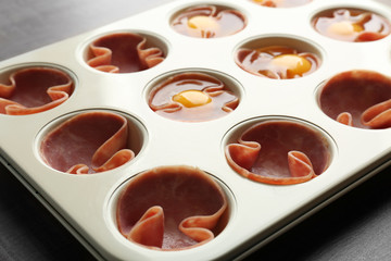 Muffin tin with raw eggs and ham on table, closeup