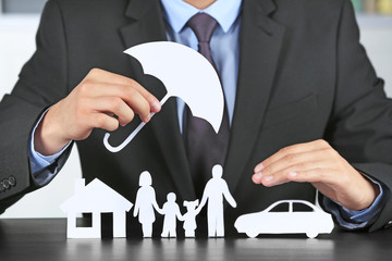 Fototapeta na wymiar Man holding umbrella over paper silhouette of family, house and car on table. Insurance concept