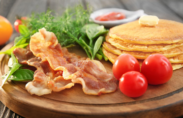 Tasty breakfast with pancakes, bacon and vegetables on wooden board, close up