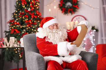 Fototapeta na wymiar Authentic Santa Claus with sheet of paper sitting in armchair at room decorated for Christmas
