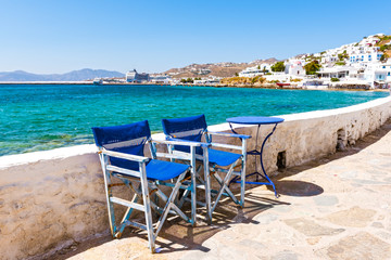 Fototapeta na wymiar Blue chairs and table on the coastal promenade with beautiful view of the sea and port in Mykonos town. Cyclades Islands, Greece.