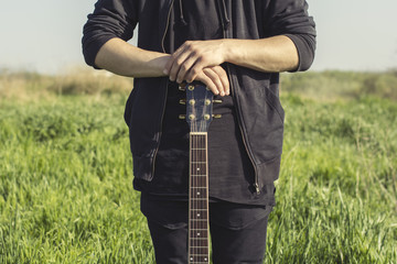 A young man in black clothes is holding a guitar with both hands against the blue sky and green grass
