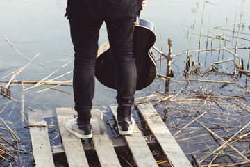 A young man in black clothes with a guitar in his hand is standing on a dilapidated bridge on the river bank