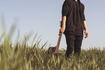A young man in black clothes is holding a guitar against the blue sky and green grass