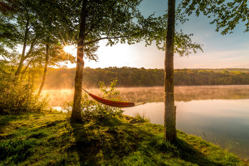 Summer camping on the lake. Empty hammock  between two trees with the view of a foggy mountain lake in sunrise light. Outdoors and adventure concept. - obrazy, fototapety, plakaty