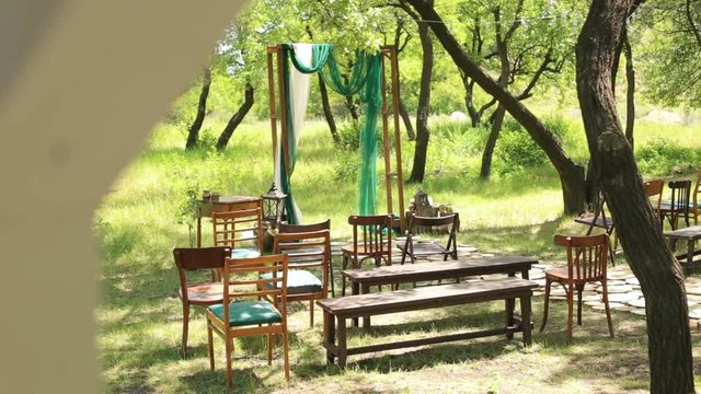 Wedding arch in rustic style with a green and white cloth with old chairs and benches in the woods.