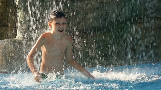 boy with a beautiful summer hot day with pleasure stands under the spray of water of the waterfall waterfall, slow motion