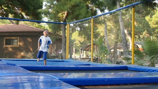 schoolboy in blue shorts and a white T-shirt with blue stripes actively jumps from the trampoline to the trampoline on the sports ground in the summer park, slow motion