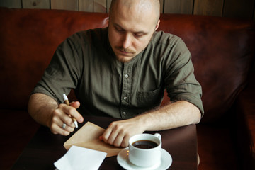 Young serious fashionable man sitting alone and writing a letter in loft-styled cafe. Cup of black coffe is standing on his table. Former factory building, natural daylight.