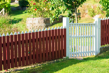 Red picket fence with white gate and garden in background.