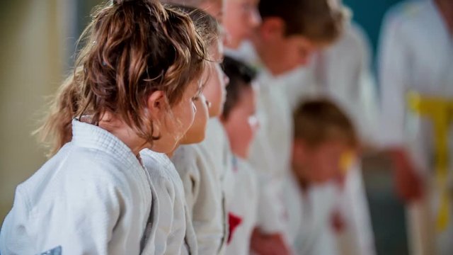 Young children are having a judo class and now, they are also sitting down on their knees. They are wearing white uniforms and having yellow and red belts on.