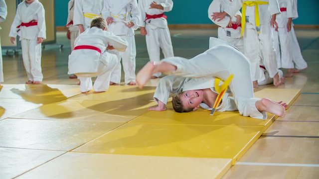 Children are doing backward somersaults. They are performing on a yellow mat in the school gym.
